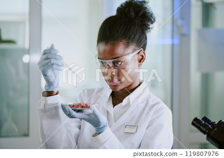 Scientist conducting research in modern laboratory while holding pipette and wearing protective gear. Focused professional carefully pipetting liquid into small container Scientist conducting research in modern laboratory while holding pipette and wearing protective gear. Focused professional carefully pipetting liquid into small container 119807976