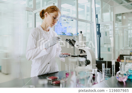 Scientist conducting research in modern laboratory with advanced equipment, surrounded by test tubes and microscope, focused on experiments involving liquids in petri dishes 119807985