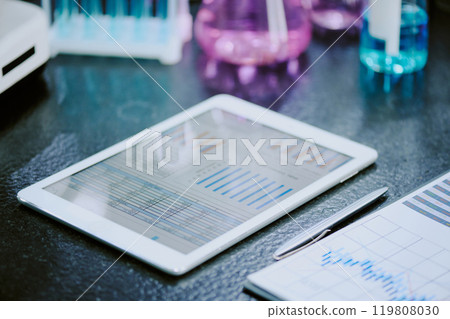 Close-up of white tablet displaying scientific graphs on lab table, surrounded by lab equipment and documents, indicating research and analysis scene 119808030