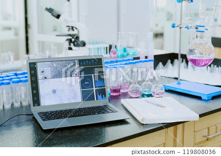 Laboratory scene featuring a laptop analyzing scientific data among various equipment and glassware, with a microscope in the background visible 119808036