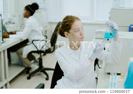 Scientist holding blue chemical solution in laboratory while a colleague working in background, both focused on tasks and using advanced equipment surrounded by scientific tools 119808037