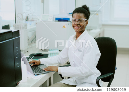 Smiling scientist facing laptop while working in laboratory, wearing protective gear. Equipment and devices in background illustrating research environment 119808039