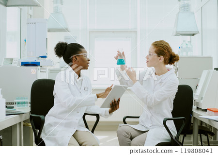 Two scientists discussing an experiment while holding a tablet and beaker in a bright laboratory with advanced equipment present, highlighting teamwork and scientific research 119808041