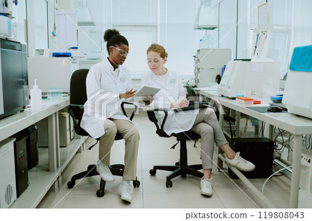 Two scientists sitting in laboratory, analyzing data on tablets while discussing research. Laboratory equipment and samples placed on counters around them Two scientists sitting in laboratory, analyzing data on tablets while discussing research. Laboratory equipment and samples placed on counters around them 119808043