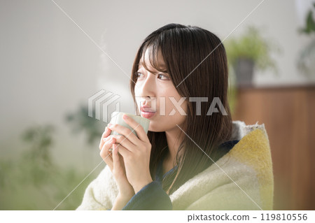 Close-up of a relaxing young woman drinking a hot drink in the living room in winter; Profile of a woman cooling down a hot drink Close-up of a relaxing young woman drinking a hot drink in the living room in winter; Profile of a woman cooling down a hot drink 119810556