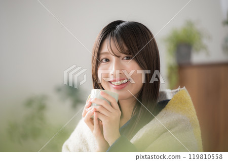 Close-up of a relaxing young woman drinking a hot drink in the living room in winter, looking at the camera 119810558