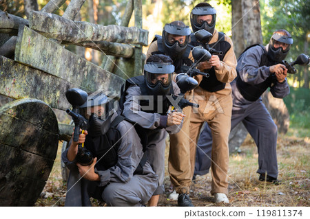 Group of paintball players aiming and shooting with guns outdoors Group of paintball players aiming and shooting with guns outdoors 119811374