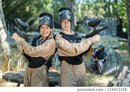 Man and woman in camouflage and masks with guns pose back to back while playing paintball outdoors 119811508