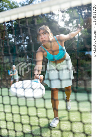 View through net of young female paddle tennis player hitting ball View through net of young female paddle tennis player hitting ball 119811720