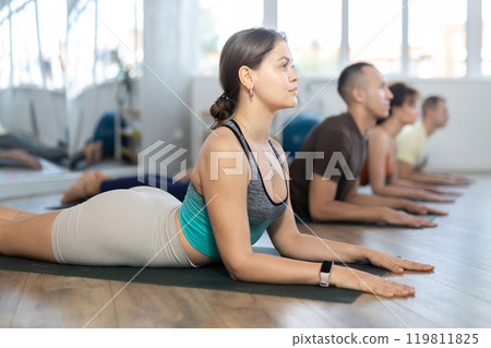 Young female doing cobra pose for health care and wellness with group together at yoga studio Young female doing cobra pose for health care and wellness with group together at yoga studio 119811825