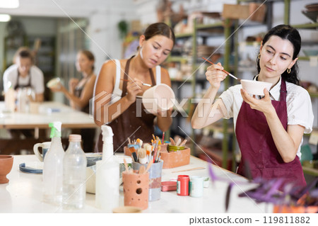 Young woman sits near table and decorates clay ceramic craft vase, plate 119811882