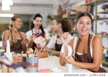 Young woman sits near table and decorates clay ceramic craft vase, plate Young woman sits near table and decorates clay ceramic craft vase, plate 119811928