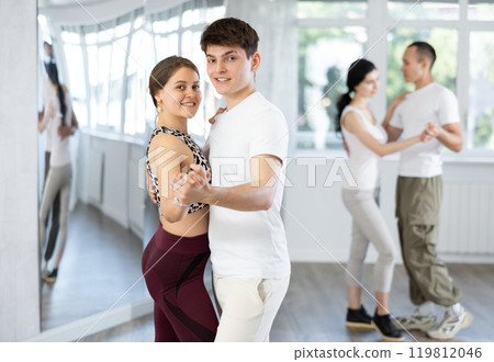 Positive young woman and man practicing waltz dance moves in pair during group training in dancing studio Positive young woman and man practicing waltz dance moves in pair during group training in dancing studio 119812046