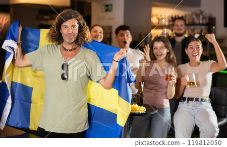 Company of sports fans waving flag of Sweden and supporting national team with beer in bar Company of sports fans waving flag of Sweden and supporting national team with beer in bar 119812050