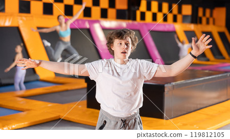 Smiling young guy in white t-shirt and gray sweatpants having great time while jumping on trampoline in game club 119812105