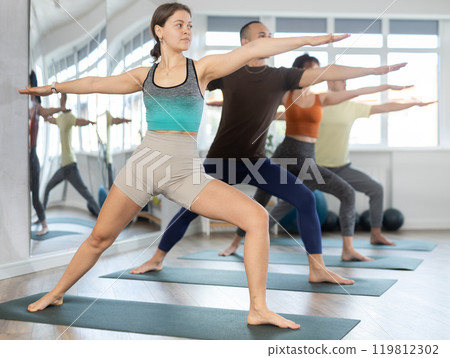 Lovers of active lifestyle on yoga mat perform doing version of warrior pose in gym Lovers of active lifestyle on yoga mat perform doing version of warrior pose in gym 119812302