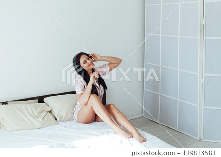 Beautiful brunette woman in pajamas listening to music in headphones in bedroom on bed Beautiful brunette woman in pajamas listening to music in headphones in bedroom on bed 119813581