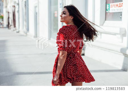 Beautiful brunette woman in a red summer dress walking through the streets of the city 119813583
