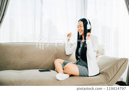 Amidst a relaxing moment on the sofa a woman holds her smartphone dances with headphones on. Immersed in music experiencing joy and entertainment at home. Concept of vibrant relaxation. Amidst a relaxing moment on the sofa a woman holds her smartphone dances with headphones on. Immersed in music experiencing joy and entertainment at home. Concept of vibrant relaxation. 119814797