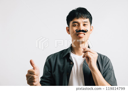 A smiling Asian man is portrayed in a portrait, holding a funny mustache card, exuding joy and humor. Isolated on white for Fathers Day and November-themed photography. 119814799