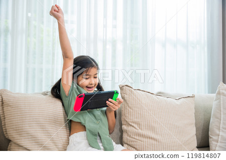 Happy Asian young girl using a tablet while sitting on a couch, excited child holding console playing video game raising hand look like win the games on sofa in living room at home online game concept Happy Asian young girl using a tablet while sitting on a couch, excited child holding console playing video game raising hand look like win the games on sofa in living room at home online game concept 119814807