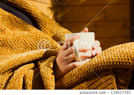 Female hands holding white cup, mug in hands. Woman wrapped in yellow plaid is drinking hot beverage - tea, cocoa, chocolate or coffee. Good morning. Autumn, winter cozy sunny day. Relax, leisure mood Female hands holding white cup, mug in hands. Woman wrapped in yellow plaid is drinking hot beverage - tea, cocoa, chocolate or coffee. Good morning. Autumn, winter cozy sunny day. Relax, leisure mood 119817391