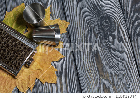 Flask for an alcoholic drink and metal glasses. Autumn maple leaves are yellow. On pine boards painted black and white. 119818384