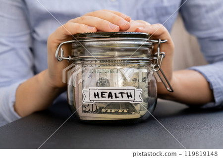Unrecognizable woman holding Glass jar full of American currency dollars cash banknote with text HEALTH. Preparation saving money. Moderate consumption and economy Unrecognizable woman holding Glass jar full of American currency dollars cash banknote with text HEALTH. Preparation saving money. Moderate consumption and economy 119819148