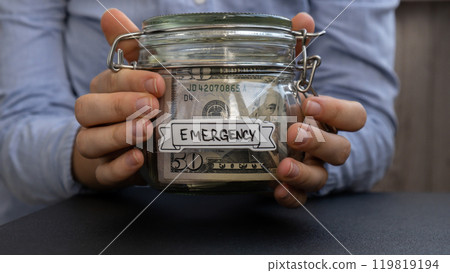 Unrecognizable woman holding Glass jar full of American currency dollars cash banknote with text EMERGENCY. Preparation saving money. Moderate consumption and economy Unrecognizable woman holding Glass jar full of American currency dollars cash banknote with text EMERGENCY. Preparation saving money. Moderate consumption and economy 119819194