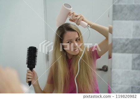 A woman is expertly styling her hair using a blow dryer and brush while in the bathroom A woman is expertly styling her hair using a blow dryer and brush while in the bathroom 119820982