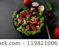 Vegetable salad of tomatoes, cucumbers, onions, lettuce dressed with aromatic dressing in a black plate on a gray background 119821006