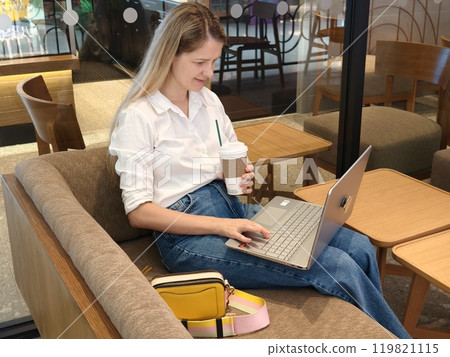 A young woman enjoys coffee while working on her laptop A young woman enjoys coffee while working on her laptop 119821115