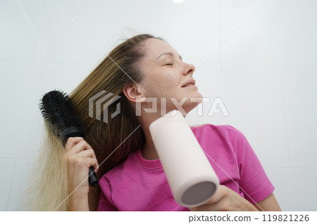 A woman is expertly styling her hair using a blow dryer and brush while in the bathroom A woman is expertly styling her hair using a blow dryer and brush while in the bathroom 119821226