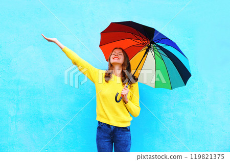 Happy smiling young woman holding colorful umbrella in autumn looking up on colorful blue background 119821375