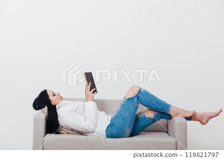 Beautiful brunette woman in lounge clothes reading a book on the sofa Beautiful brunette woman in lounge clothes reading a book on the sofa 119821797