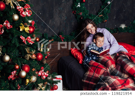 Mom and son reading a book at the Christmas tree with gifts for the new year 119821954