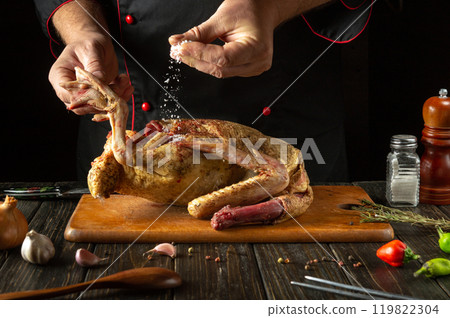 Hands sprinkle salt over a seasoned whole duck, highlighting the culinary artistry. Fresh herbs and spices surround a wooden cutting board, creating a cozy hotel kitchen atmosphere Hands sprinkle salt over a seasoned whole duck, highlighting the culinary artistry. Fresh herbs and spices surround a wooden cutting board, creating a cozy hotel kitchen atmosphere 119822304