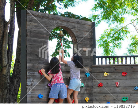 Children play outdoors on a wooden playground in a park. Happy girl playing on wooden climbing wall in playground during summer. 119822719