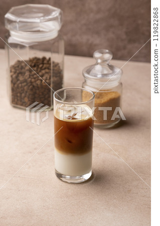 Coffee with coconut milk and ice cubes in a tall glass. Traditional non-alcoholic cold drink. In the background, a jar of coffee and a jar of sugar. Selective focus, close-up. Coffee with coconut milk and ice cubes in a tall glass. Traditional non-alcoholic cold drink. In the background, a jar of coffee and a jar of sugar. Selective focus, close-up. 119822868
