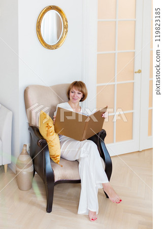 An adult beautiful woman spends time in her stylish living room and holds a big book in her hands. An adult beautiful woman spends time in her stylish living room and holds a big book in her hands. 119823185