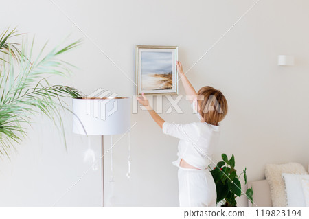 Woman hanging picture on wall in room. Interior design. Woman hanging picture on wall in room. Interior design. 119823194