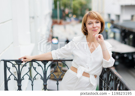 Adult woman in white suit is standing on balcony in town and looking at camera. Adult woman in white suit is standing on balcony in town and looking at camera. 119823195