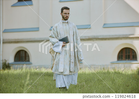 Young Caucasian priest in golden white robe holding Bible in hands while walking through yard Young Caucasian priest in golden white robe holding Bible in hands while walking through yard 119824618