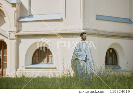 Caucasian priest in golden white robe walking slowly through yard with fresh cut grass Caucasian priest in golden white robe walking slowly through yard with fresh cut grass 119824619