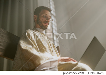 Low angle shot of young priest typing on keyboard while sitting on bench in church 119824628