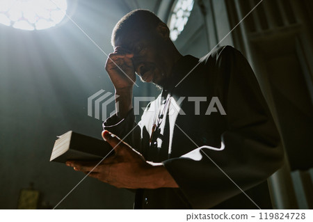 Low angle shot of African American priest in robe holding closed Bible crossing himself during church service 119824728