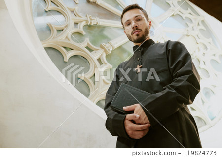 Low angle portrait of young Caucasian priest holding Bible looking at camera while standing next to beautiful and majestic white wall of church 119824773