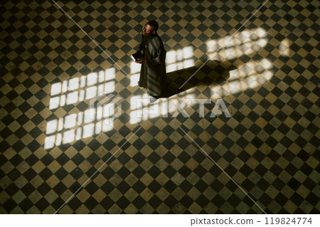 Aerial view of African American priest in black robe with holy book looking at huge vertical window while standing in nave 119824774