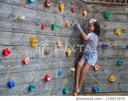 Children play outdoors on a wooden playground in a park. Happy girl playing on wooden climbing wall in playground during summer. Children play outdoors on a wooden playground in a park. Happy girl playing on wooden climbing wall in playground during summer. 119825970
