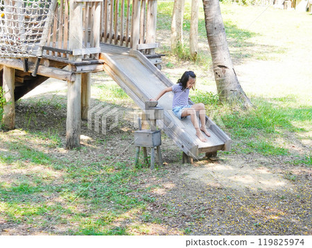 Children play outdoors on a wooden playground in a park. Happy girl playing on slide in playground in summer. 119825974